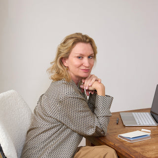 A woman with blonde hair sits in a white chair at a wooden desk, wearing a patterned beige and black blazer and brown pants. A laptop and notebook are on the desk in front of her against a plain light gray wall.